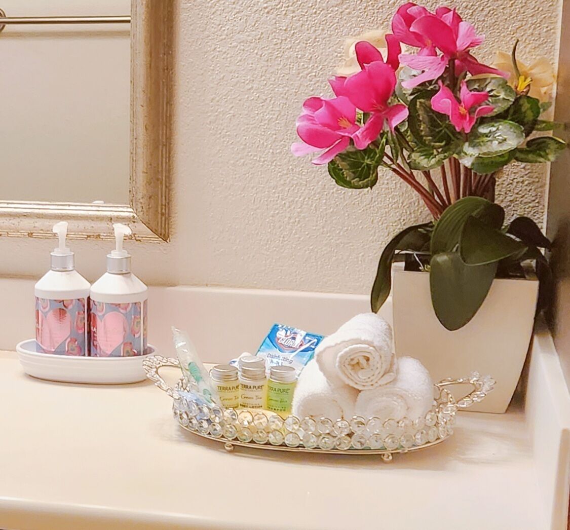 A bathroom vanity with a mirror, two soap dispensers, a tray with toiletries, a rolled towel, and a potted plant.