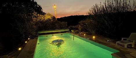Ein Schwimmbecken mit Whirlpool, umgeben von einer Sonnenterrasse mit Stühlen und einem Sonnenuntergang im Hintergrund.