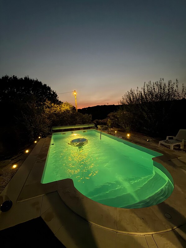 A swimming pool with a hot tub, surrounded by a deck with chairs and a sunset in the background.