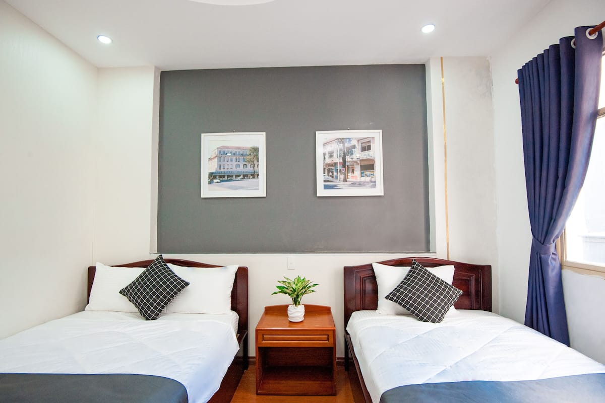 A hotel room with two single beds, a small wooden nightstand with a plant, and two framed pictures on the wall.