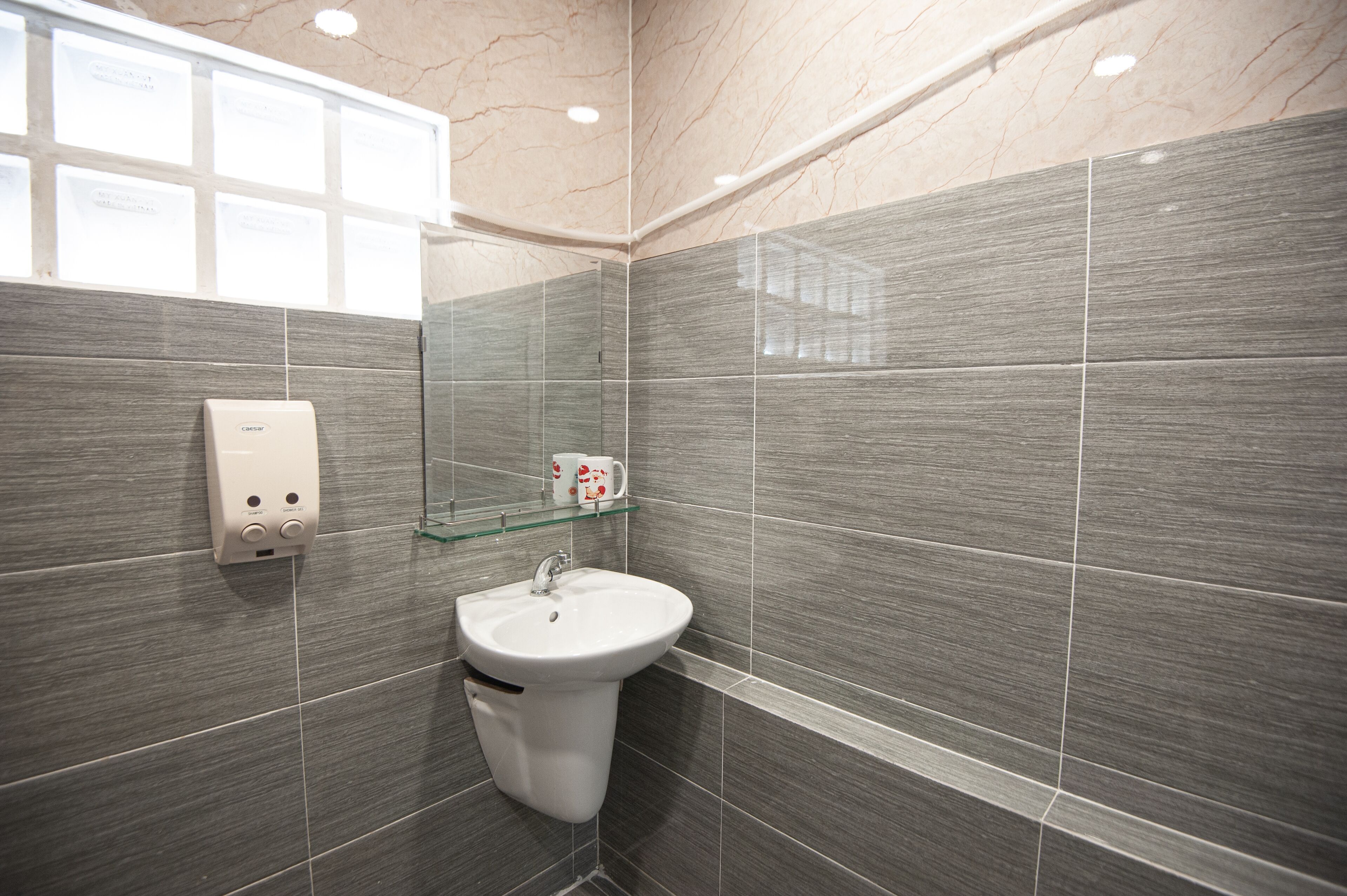 A modern bathroom with a grey tiled wall, a white sink, and a glass shelf with toiletries.