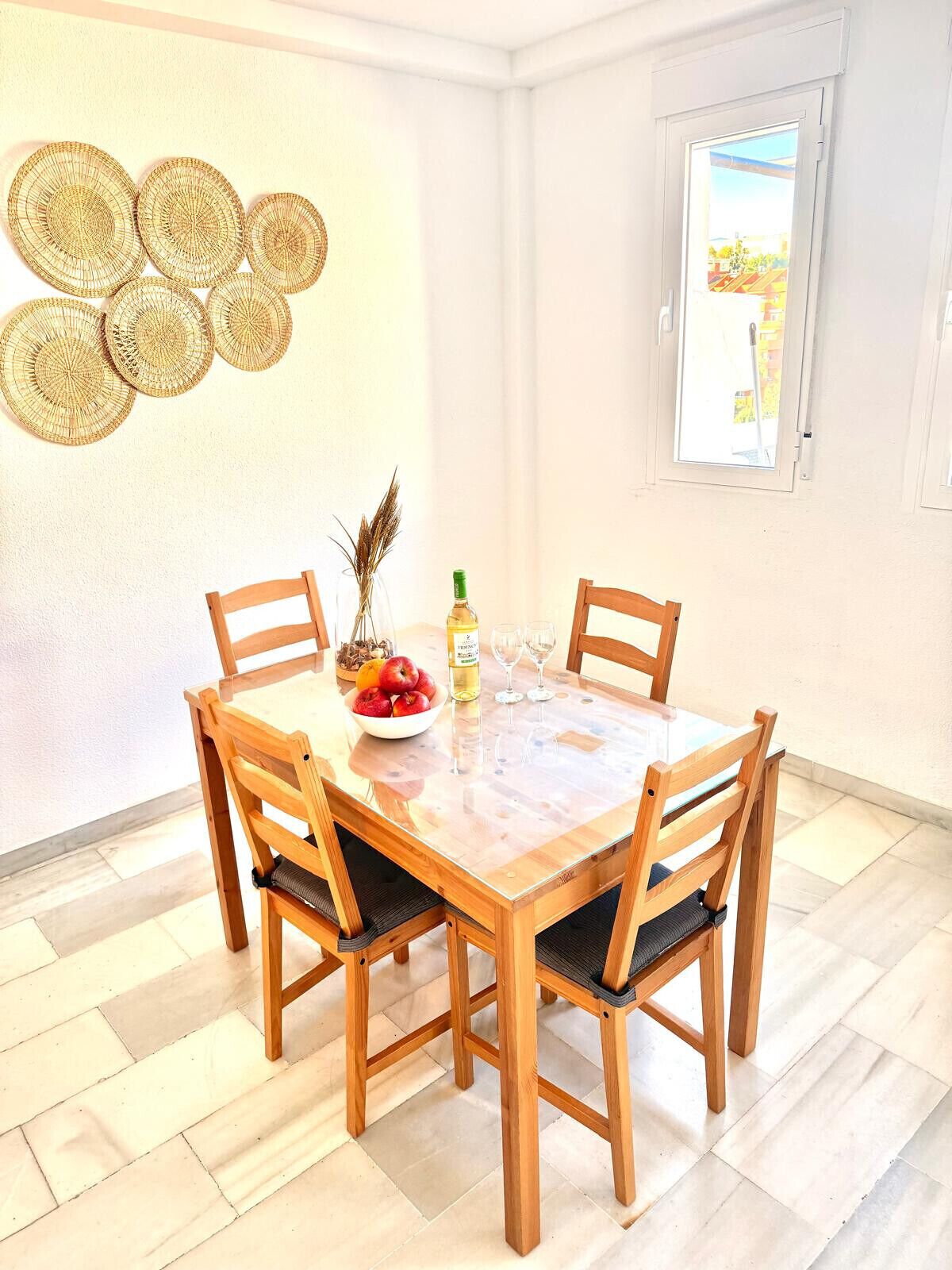 A dining area with a round wooden table and four chairs, a bowl of fruit, a bottle of wine, and a vase with dried plants.