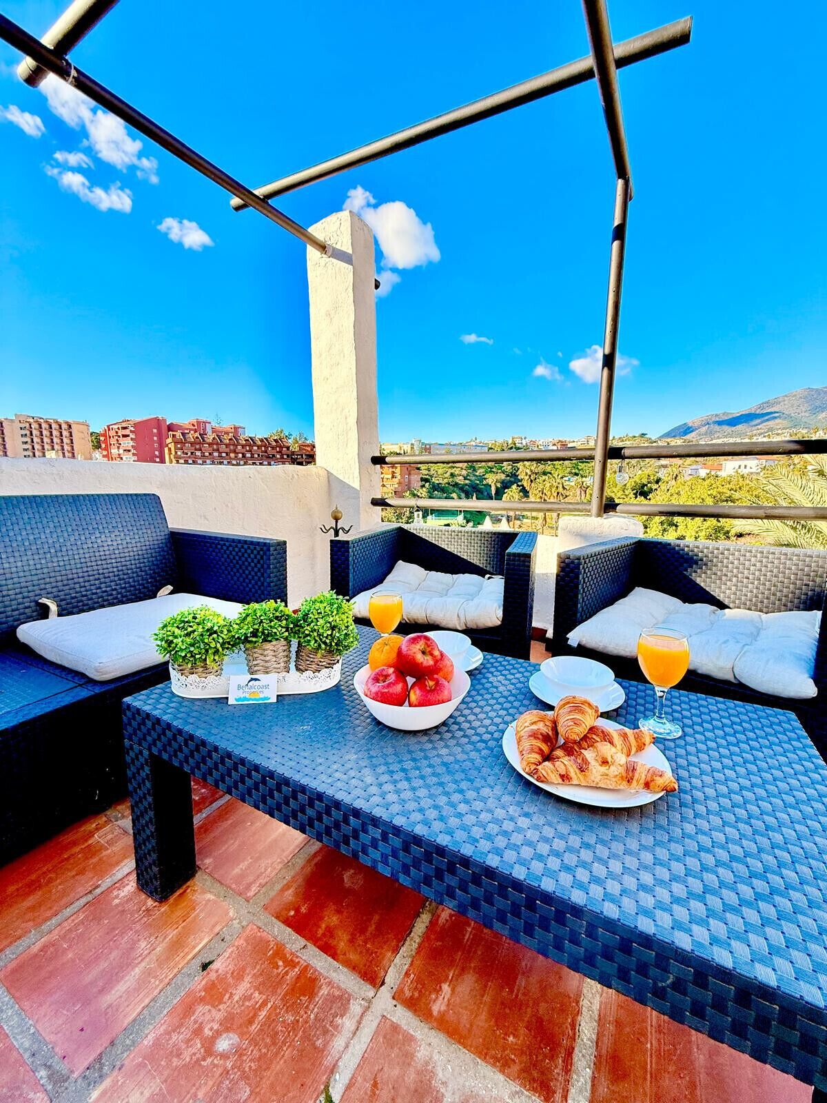 Una terraza en la azotea con mobiliario exterior, una mesa dispuesta con frutas, cruasanes y jugo de naranja, y una vista de la ciudad y las colinas al fondo.
