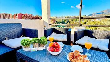Una terraza en la azotea con mobiliario exterior, una mesa dispuesta con frutas, cruasanes y jugo de naranja, y una vista de la ciudad y las colinas al fondo.
