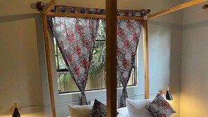 A room with a four-poster bed, a small side table with a lamp, and a window with patterned curtains. - Tanzanin Eco Home & Camp (Arusha)