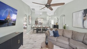 A spacious living room with a high ceiling fan, a large TV, and a modern dining area.