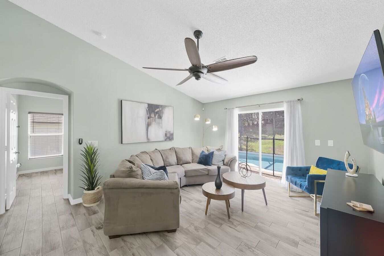 A spacious living room with a sofa, a coffee table, and a ceiling fan. There is a large window with curtains and a view of a pool.