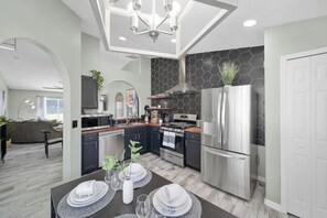 A modern kitchen with stainless steel appliances, a dining area with a table set for four, and a living room with a sofa and a TV. - 8052 Heated Pool Grill House Kissimmee by Disney (Kissimmee)