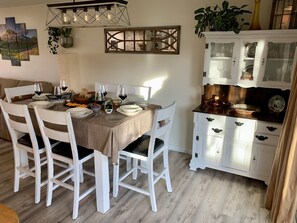 Comedor con una mesa dispuesta para cuatro personas, rodeada de sillas blancas. A la derecha, un mueble con puertas de vidrio.