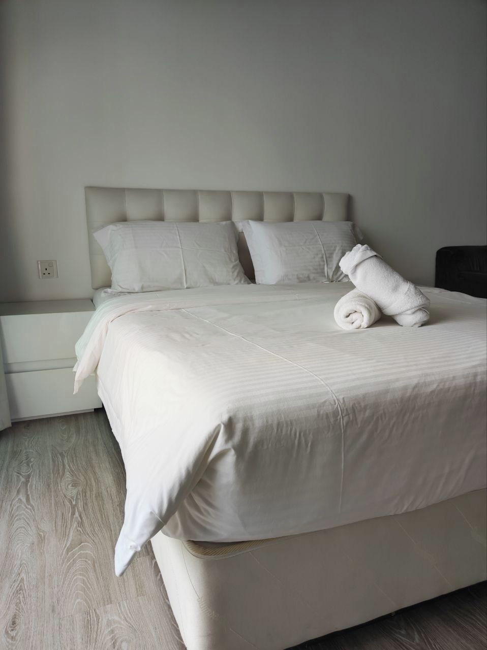 A neatly made bed with a rolled towel, a headboard, and a bedside table with a lamp.