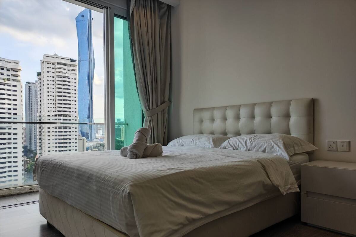A modern hotel room with a large bed, a nightstand, and a view of tall buildings through a large window.