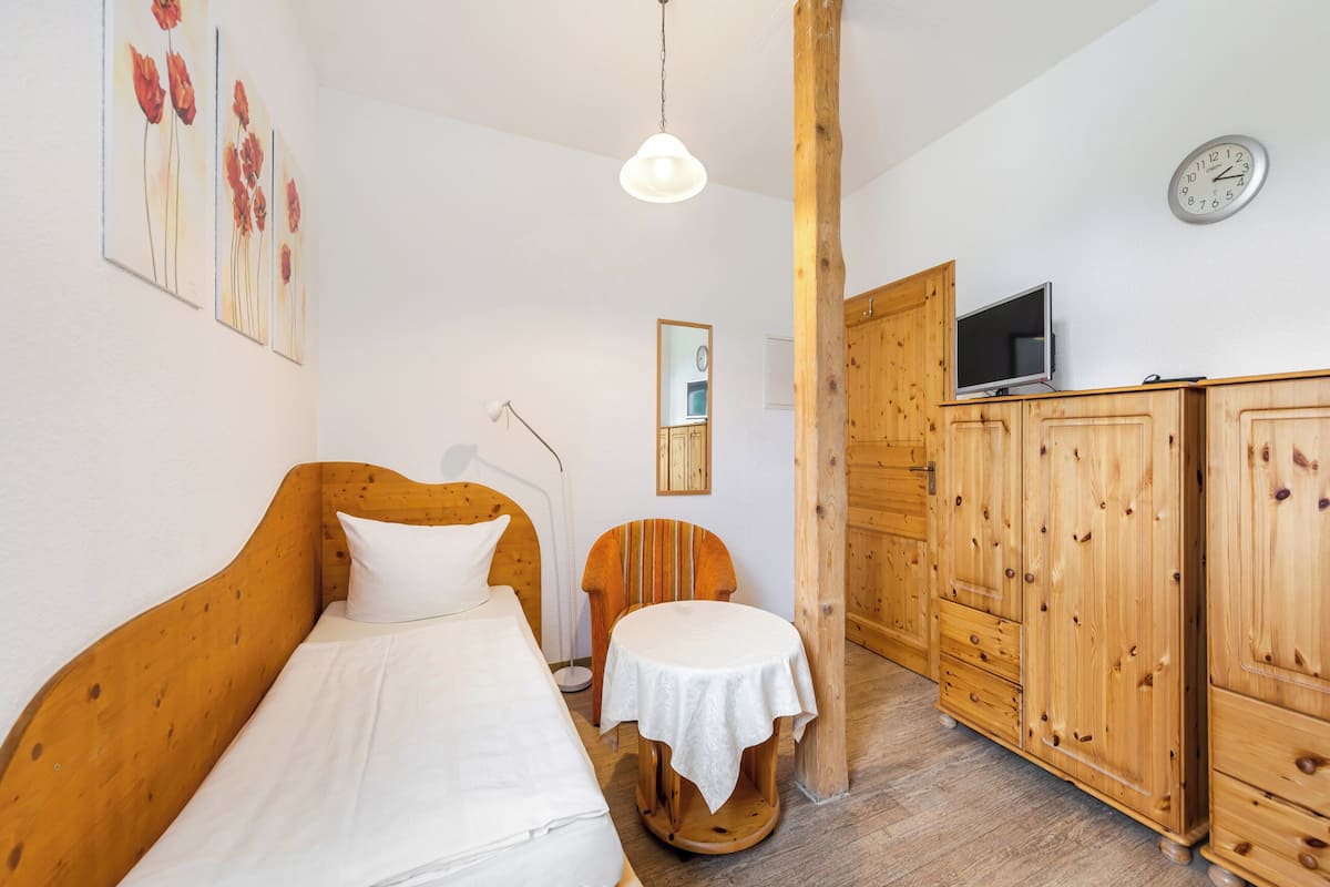 Une chambre à coucher avec un lit en bois, une petite table ronde recouverte d’une nappe blanche, une chaise en bois, un téléviseur posé sur une commode en bois et une horloge murale.