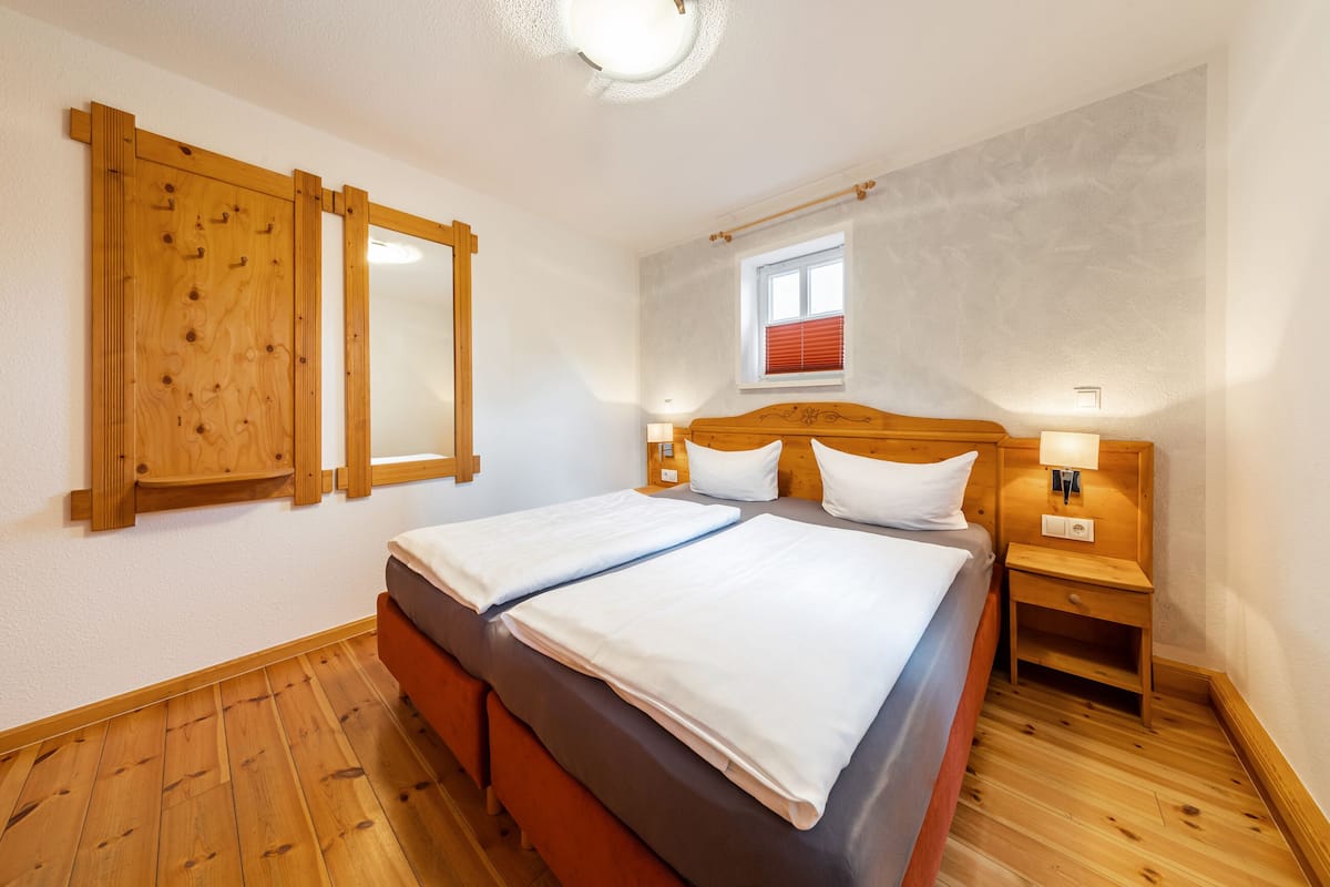 Un lit double avec du linge de lit blanc, une tête de lit en bois et une table de chevet avec une lampe. On y trouve également une armoire en bois avec un miroir, une fenêtre avec des stores et un luminaire au plafond.