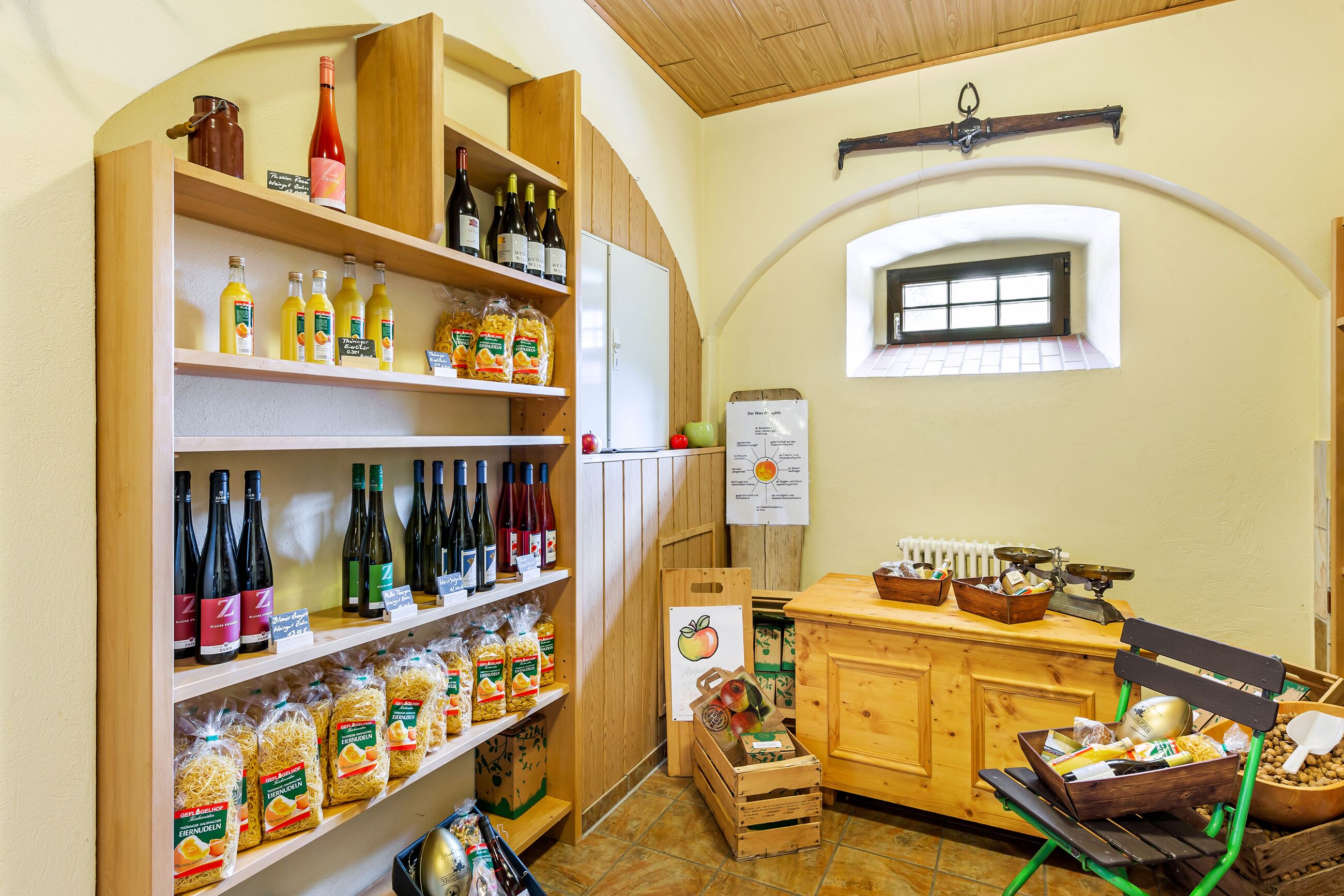 A kitchen with wooden shelves stocked with wine bottles and pasta, a wooden counter with a scale, and a window with a view of the outside.