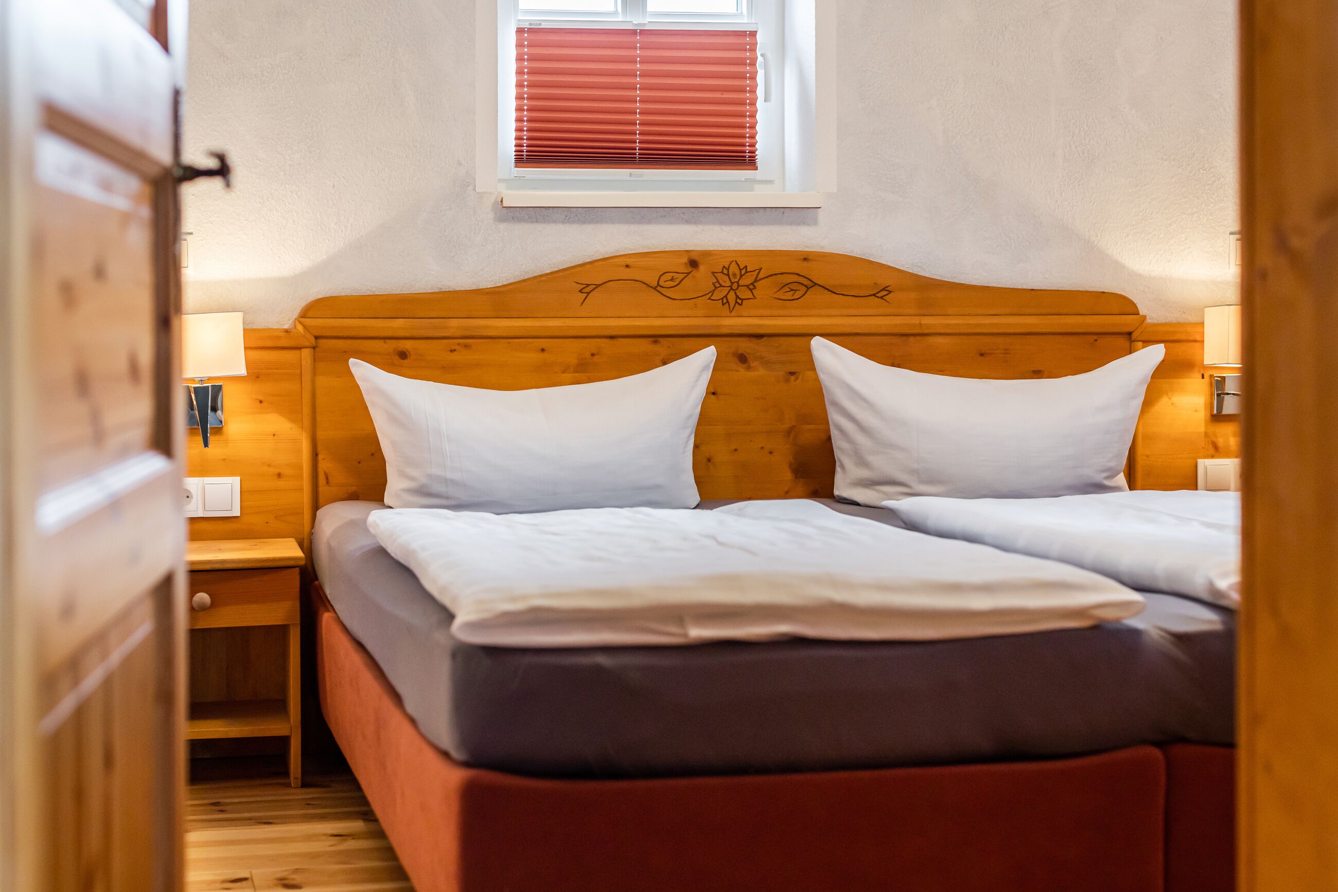A double bed with white linens, a wooden headboard with floral carvings, a nightstand with a lamp, and a window with red blinds.