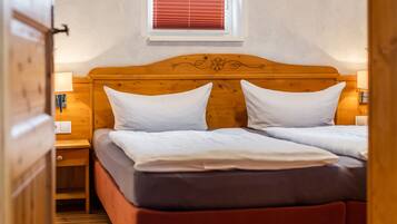 A double bed with white linens, a wooden headboard with floral carvings, a nightstand with a lamp, and a window with red blinds.