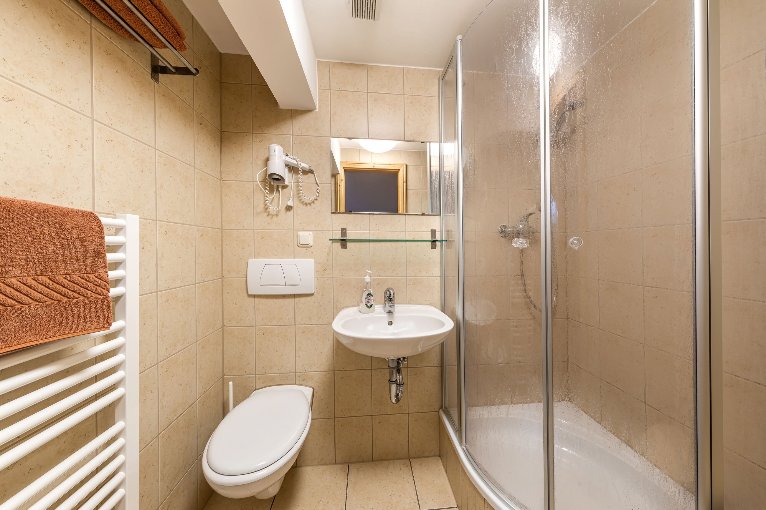 Une salle de bain avec des toilettes blanches, des carreaux beiges, une douche en verre et un lavabo blanc avec un miroir au-dessus.