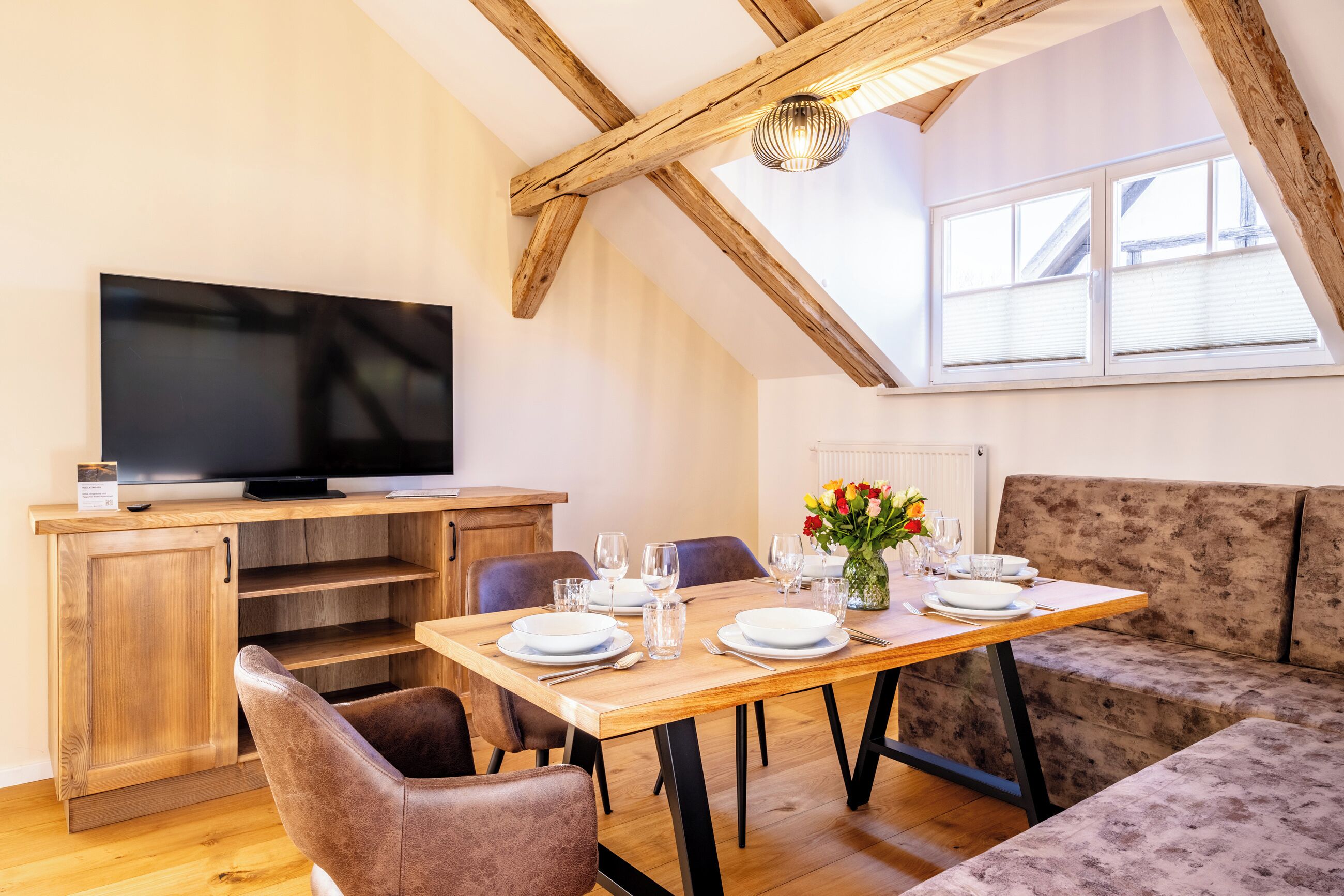 A dining area with a wooden table set for four, a television on a wooden cabinet, and a sofa.