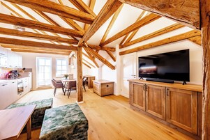 Uma sala de estar ampla com vigas de madeira aparente, uma TV de tela grande, armários de madeira e uma área de jantar com mesa e cadeiras.