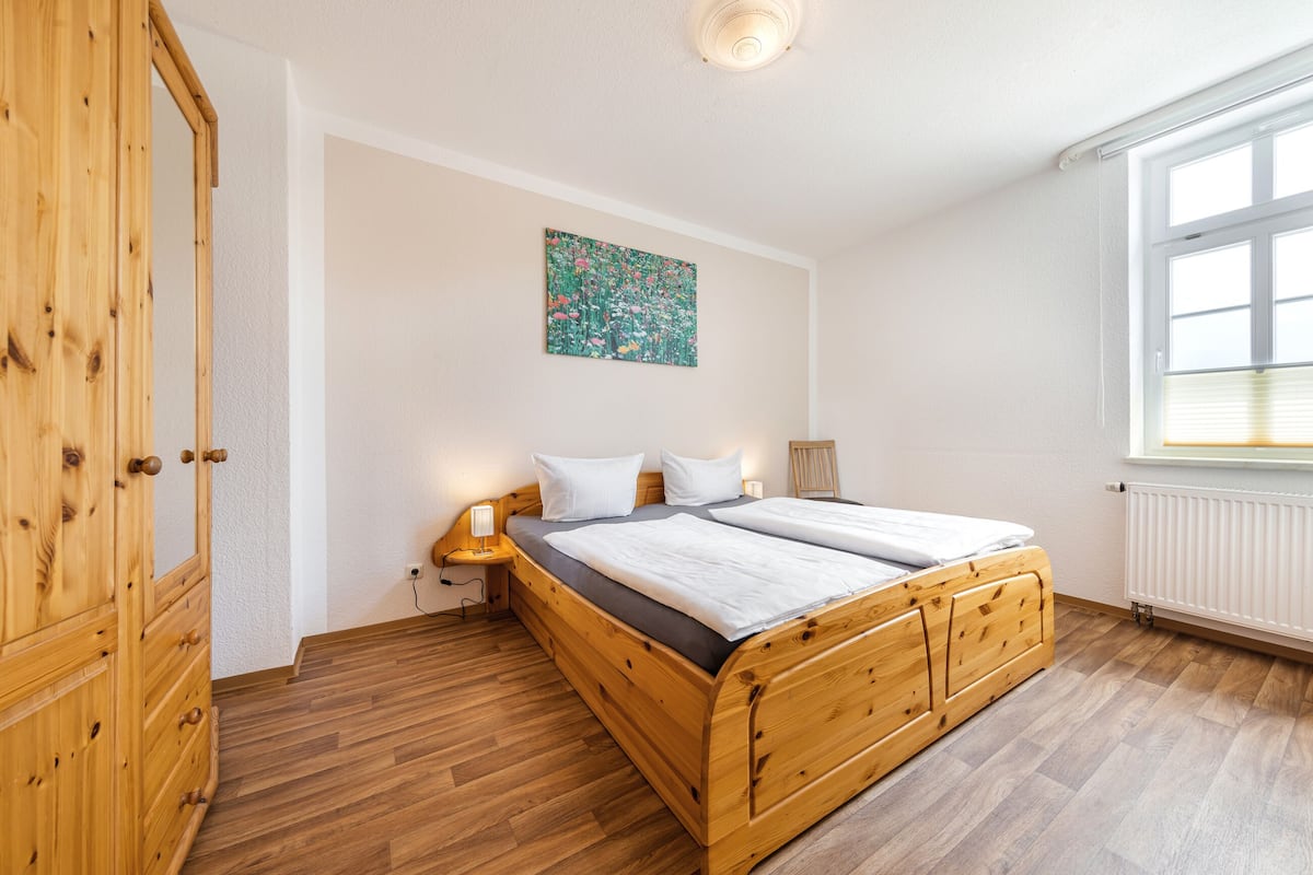 Une chambre à coucher avec un lit en bois, une table de chevet, un tableau accroché au mur et une fenêtre avec des stores.