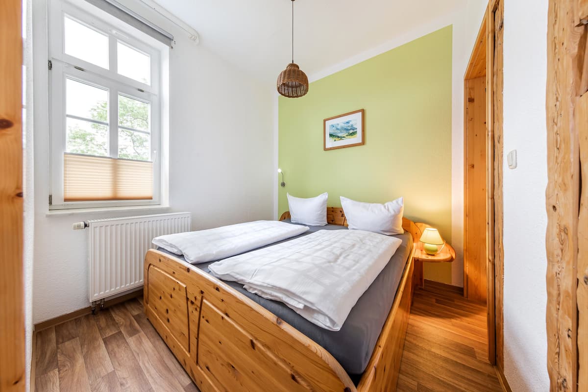 Une chambre à coucher avec un lit double, une fenêtre avec des stores, une table de chevet avec une lampe et un tableau encadré au mur.