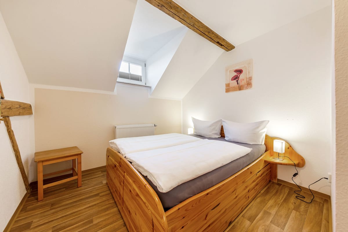 Une chambre avec un plafond en pente, un lit en bois, une table de chevet, une lampe et une petite fenêtre.