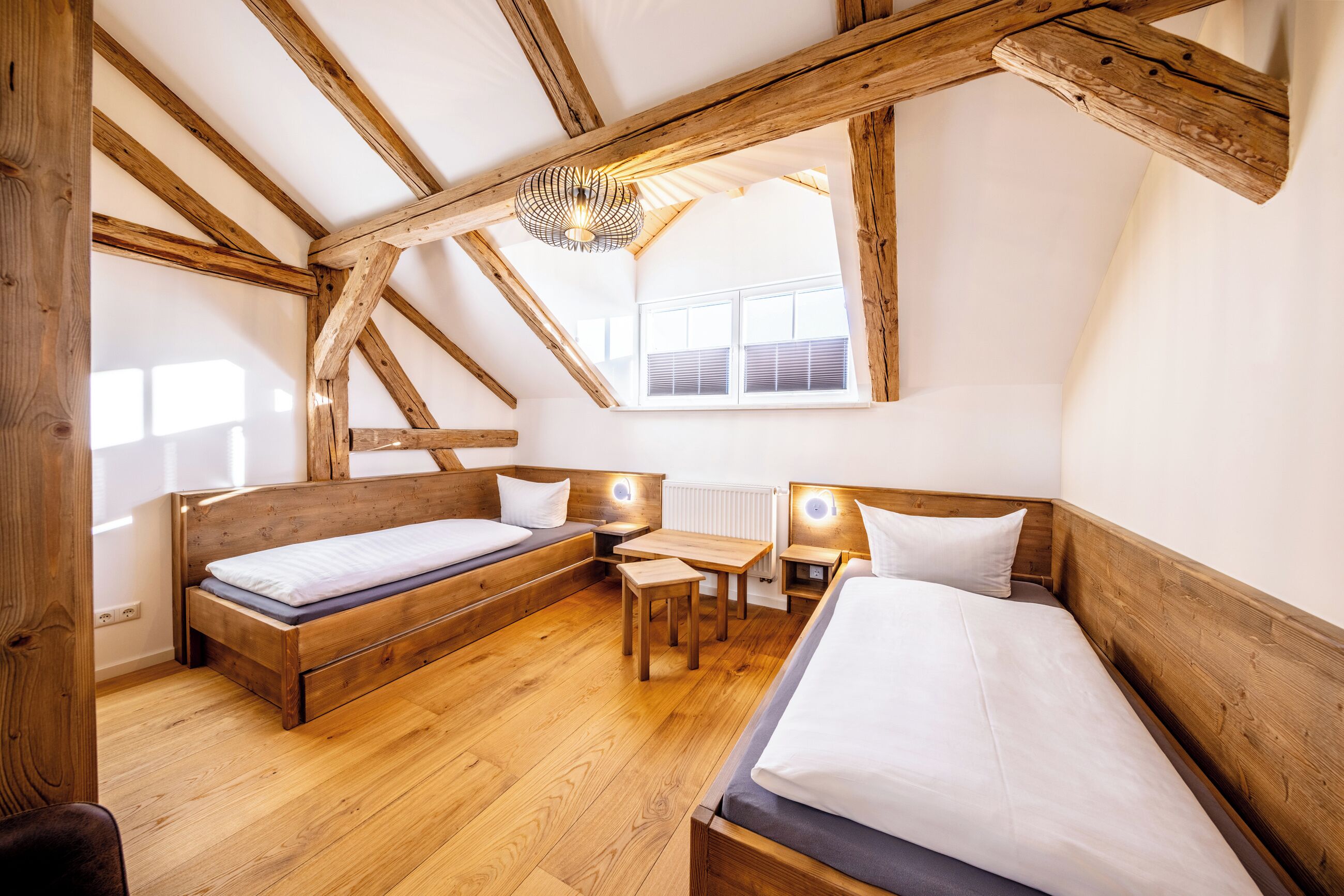 Ein gemütliches Schlafzimmer im Dachgeschoss mit Holzbalken, einem Schlafplatz zur erhöhten Etage, einem Schreibtisch und einem Fenster, das für natürliches Licht sorgt.