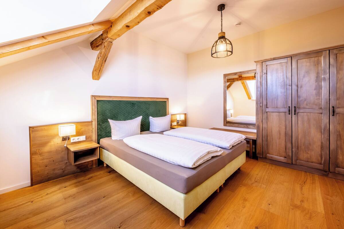Une chambre avec un plafond en bois, un lit recouvert de draps blancs, une table de chevet, un miroir et une armoire.