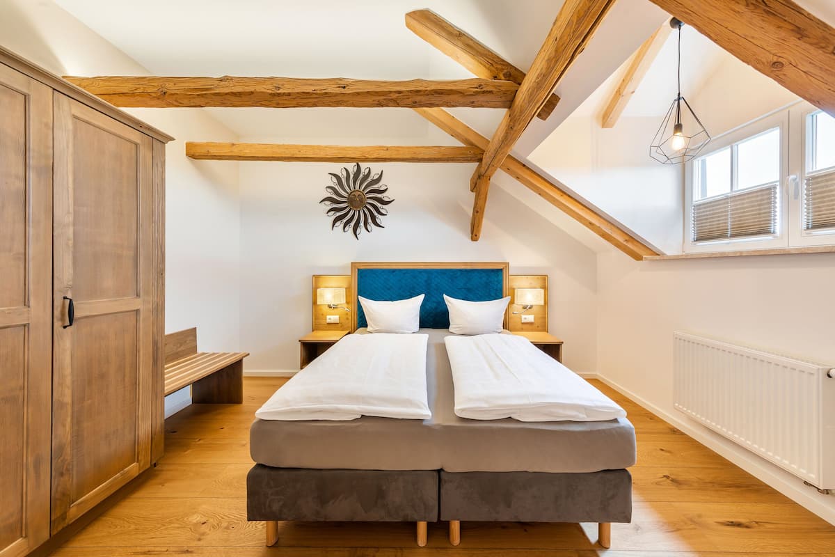 Une chambre avec un plafond en bois, un lit recouvert de draps blancs, deux tables de chevet avec des lampes et une fenêtre avec des stores.
