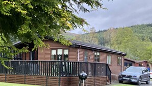 A wooden cabin with a fenced yard, a barbecue grill, and a parked car.
