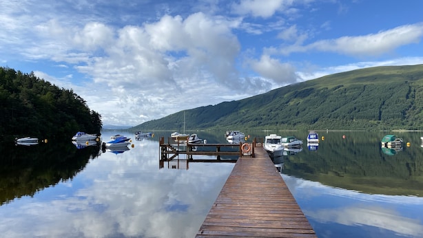 A wooden pier extending into calm water with boats docked on both sides, surrounded by forested hills.