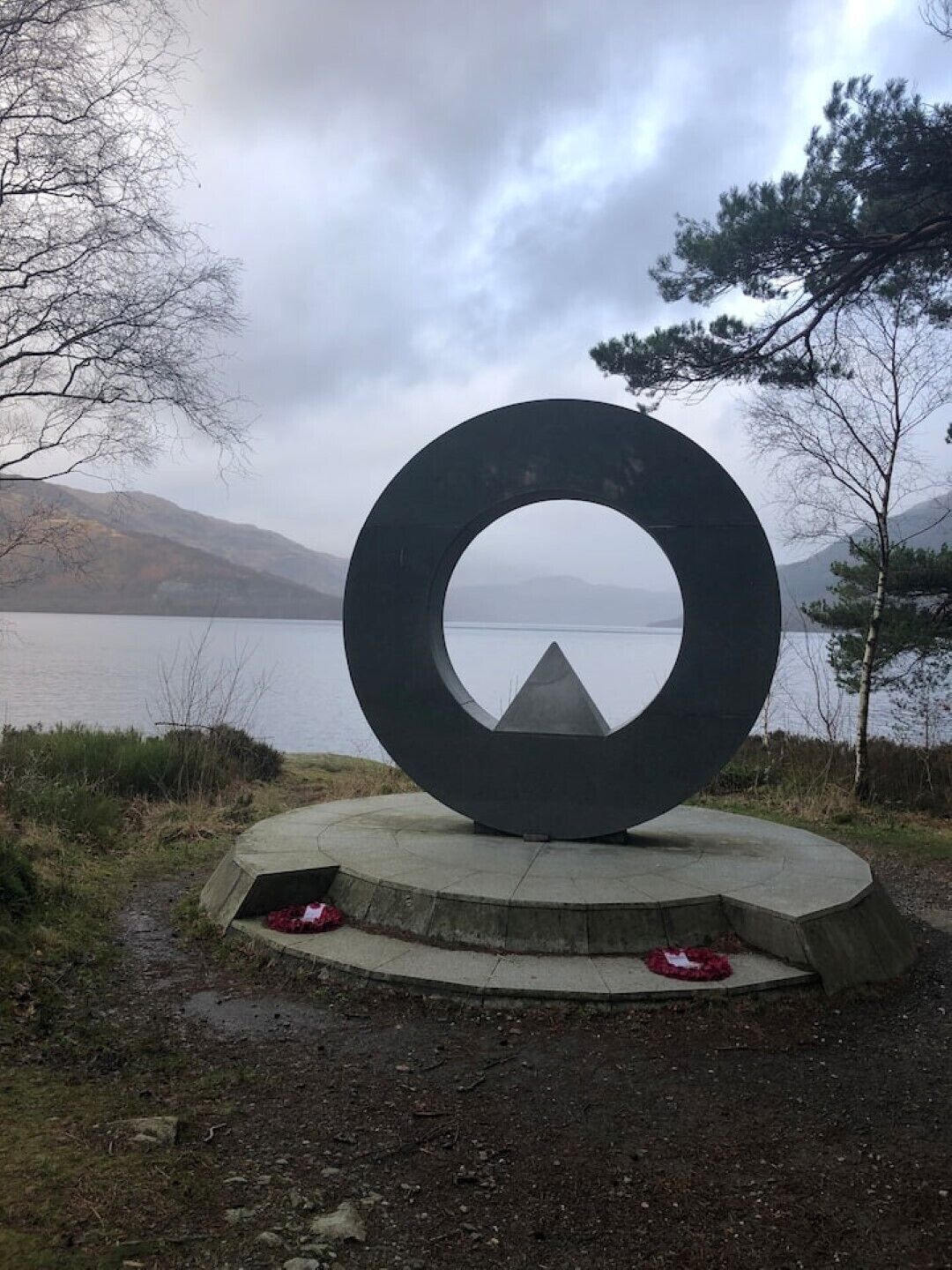 Une sculpture circulaire, dont le point central est entouré d’une base en béton, se dressant sur un fond composé d’un lac et de montagnes.