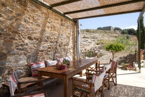 An outdoor dining area with stone walls, wooden furniture, and a view of a hill with a building in the distance. - Agritourism house in organic farm 1. (Heraklio Creta)