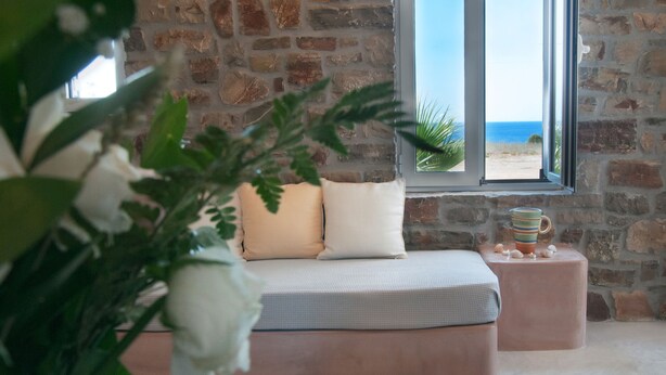 A room with a stone wall, a large window with a view of the beach, a bed with pillows, and a side table with a mug. - ANEMES (Kithira)