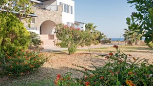 A modern building with a stone facade, an arched entrance, and a balcony overlooking a landscaped garden with various plants and flowers. - ANEMES (Kithira)