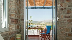A room with stone walls, a wooden table, a blue chair, a hat, and a view of the sea through a glass door. - ANEMES (Kithira)