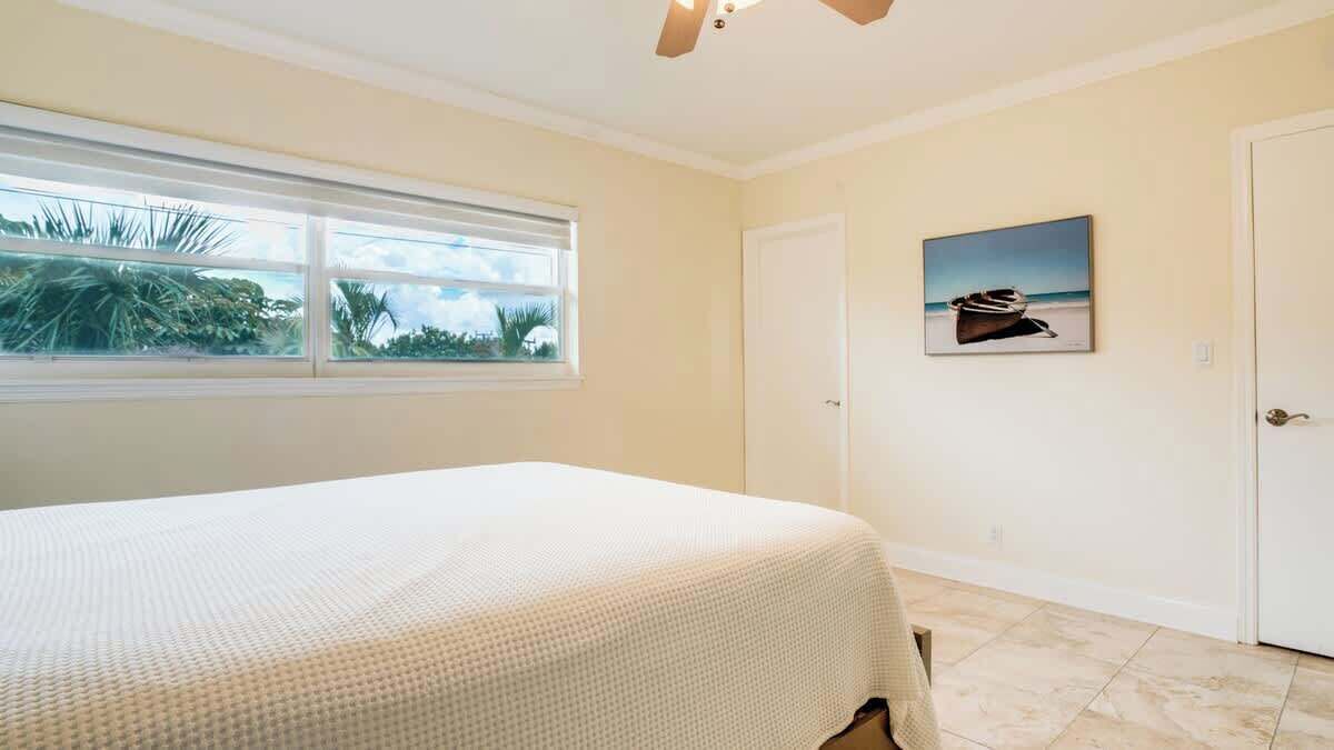 Een slaapkamer met een bed, een raam met uitzicht op palmbomen, een deur en een schilderij aan de muur.