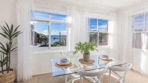 A dining area with a glass table, white chairs, and a view of the sea through large windows. - Luxe-Coastal Balmoral Beachfront (Mosman)