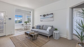 A living room with a grey sofa, a coffee table, a painting of a beach scene, and a view of the outdoors. - Luxe-Coastal Balmoral Beachfront (Mosman)