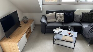 Una sala moderna con una televisión de pantalla plana, un mueble de madera, una mesa de centro de vidrio y un sofá de cuero negro con cojines estampados.