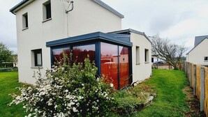 Een modern huis met twee verdiepingen en een grote glazen conservatorium, omgeven door een verzorgd gazon en een bloeiende struik.