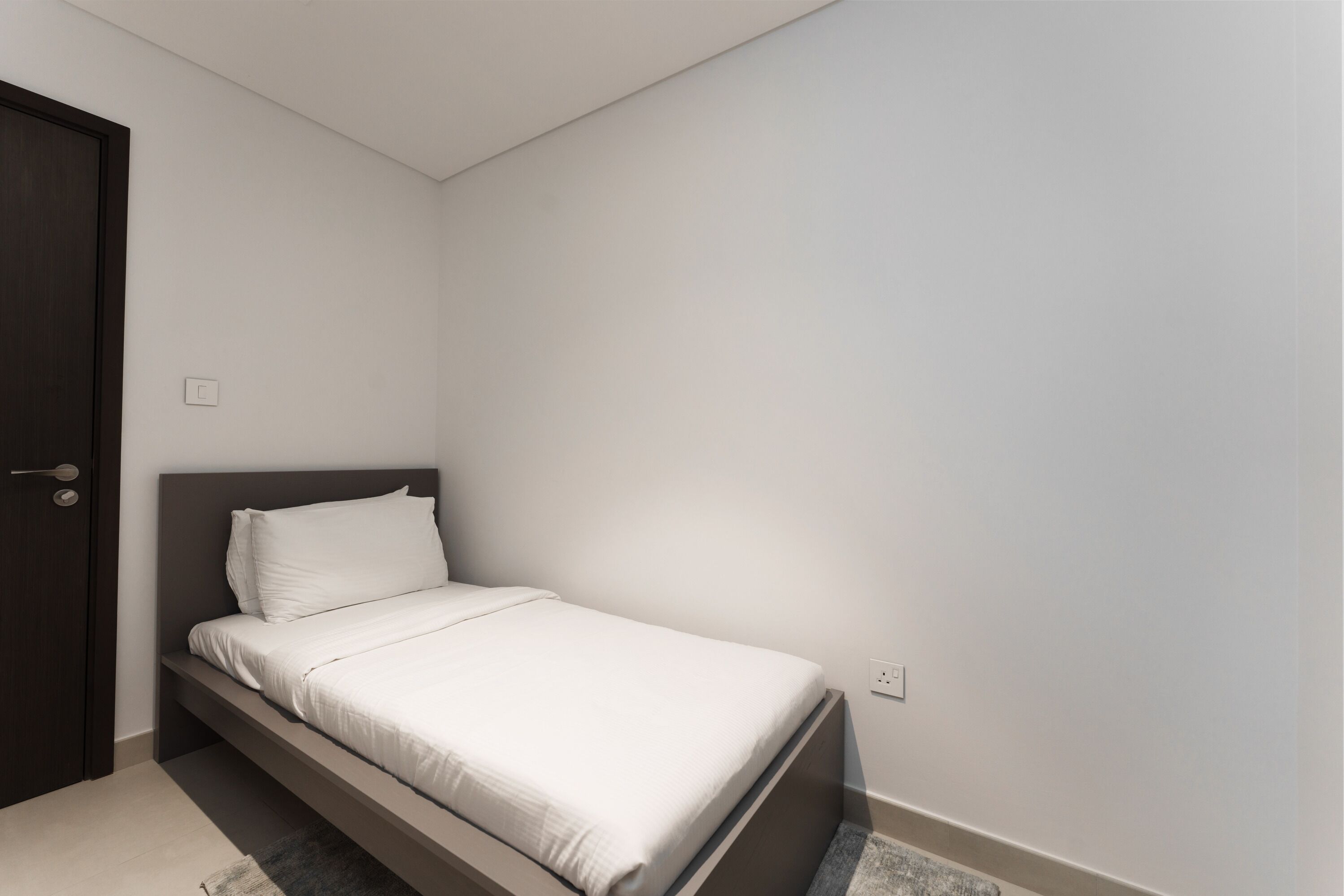 Ein kleines, ordentlich bezogenes Bett in einem schlichten Zimmer mit einer dunklen Tür und hellen Wänden.