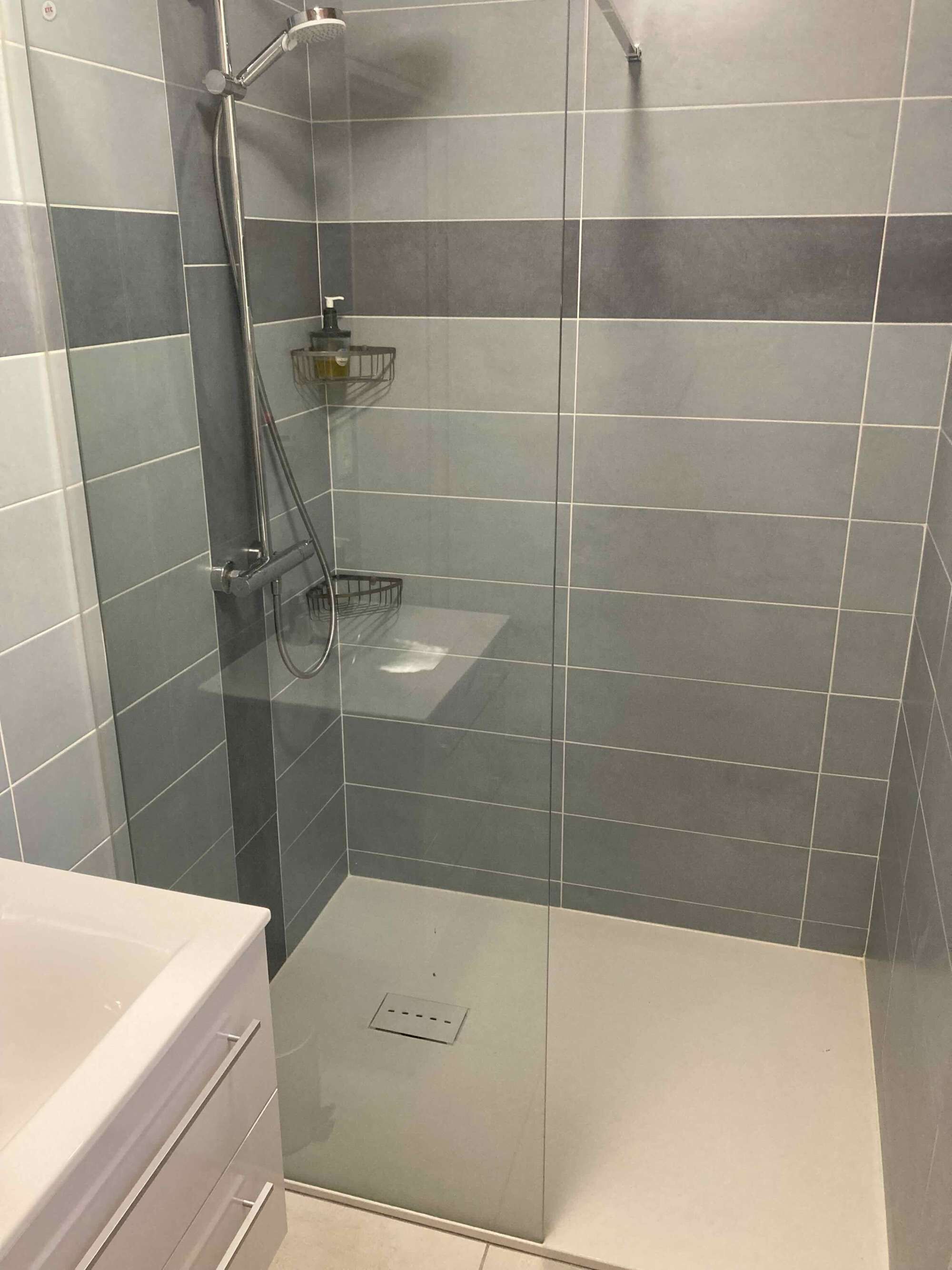 A modern shower with a glass door, grey tiles, and a white sink.