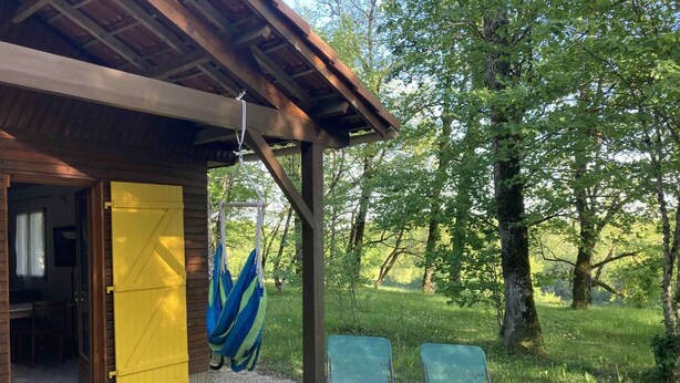 Una cabaña de madera con una puerta amarilla, una hamaca y dos sillas verdes afuera.