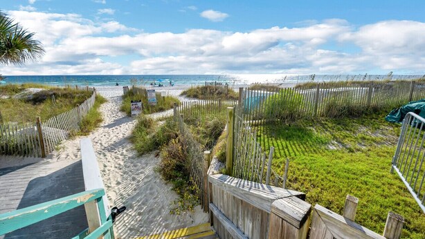 Un sentier qui longe la plage et mène à l’océan, bordé de clôtures en bois et de végétation.