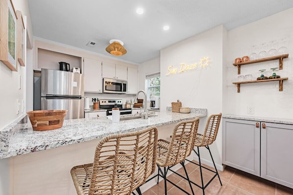A modern kitchen with a central island, stainless steel appliances, and wicker bar stools. - SUN-DAZE | Renovated 3bed Home & WALK TO BEACH! (Panama City Beach)