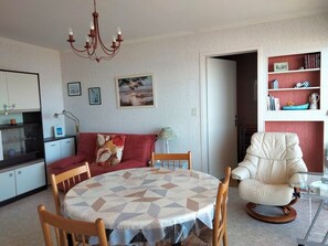 A dining area with a round table, chairs, a chandelier, a sofa, and a bookshelf. - 2-Room Apartment with Balcony and Parking - Les Sables-d'Olonne (Les Sables-d'Olonne)