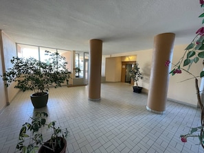 A spacious indoor area with tiled flooring, large columns, and potted plants.