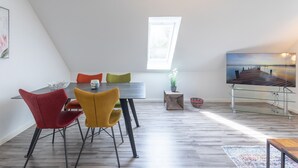 A modern dining area with a high ceiling and skylight, featuring a wooden table, colorful chairs, and a view of a dock through a window. - Vacation apartment Achterdeck in Drelsdorf for up to 4 people (Drelsdorf)