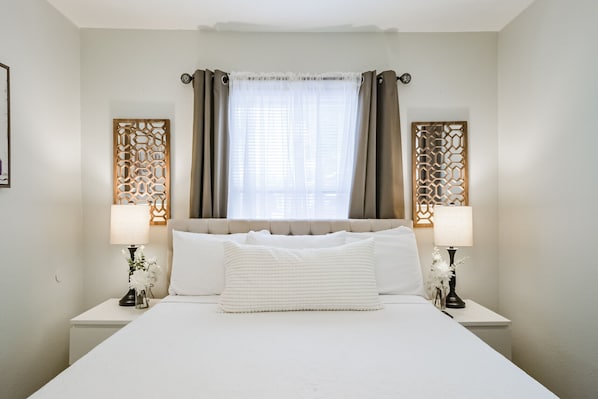 A neatly arranged hotel room with a large bed, two bedside tables with lamps, and decorative panels on the walls. - Charming private home sleeps 6 in the Heights near Downtown with fast WiFi, smart TVs, ample parking, and a primary king bed for a comfortable stay. (Houston)